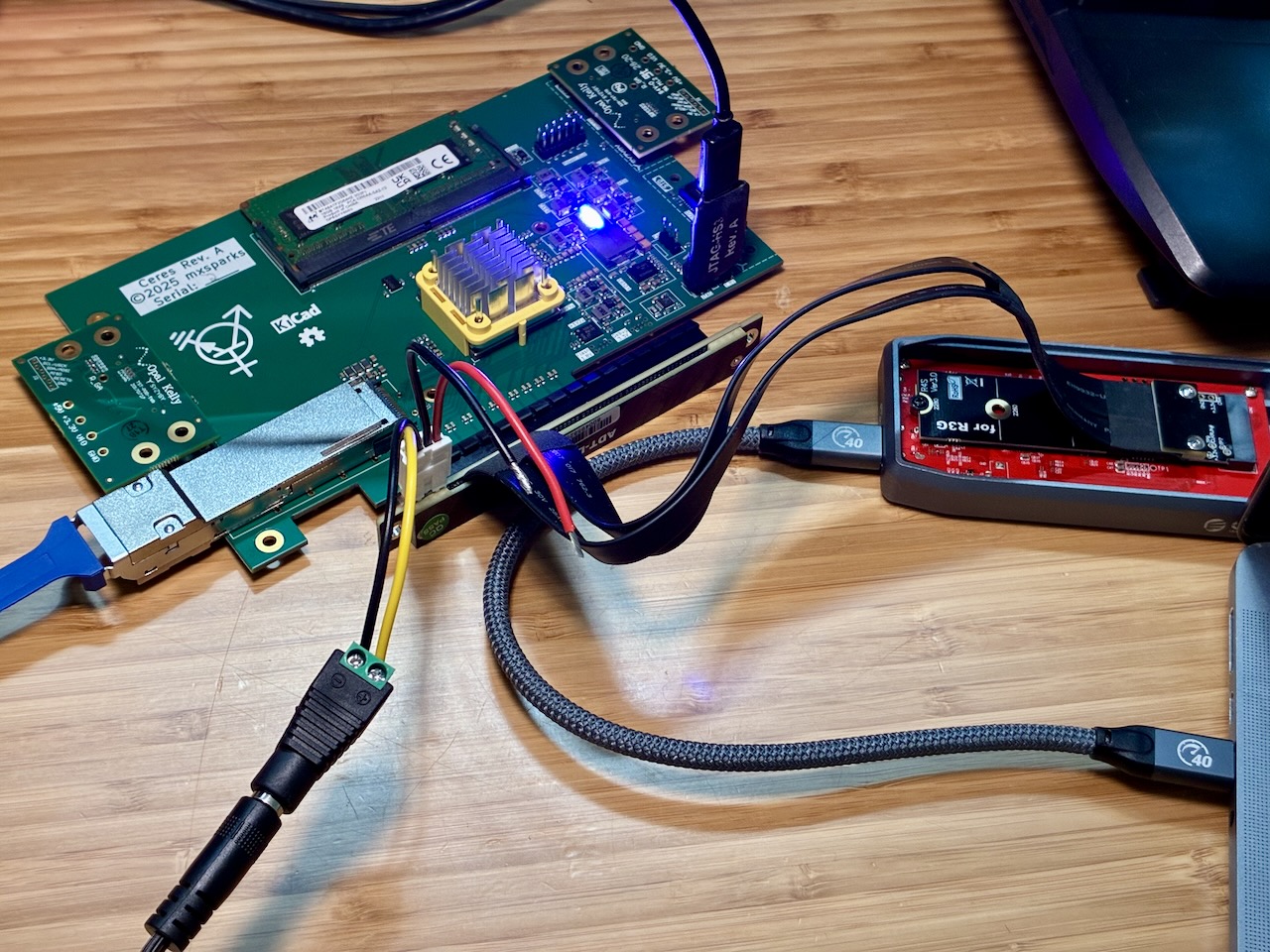 A PCI Express card sitting on a wooden table, with a DDR4 DRAM stick,
a QSFP28 loopback module, two half-length SYZYGY modules, and a JTAG
connector plugged into it. The card edge is plugged into a PCIe-to-M.2
adapter, which is in turn connected to a DC power supply and an
M.2-to-Thunderbolt adapter.